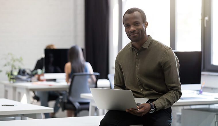 Black man sitting on the corner of his office desk with laptop