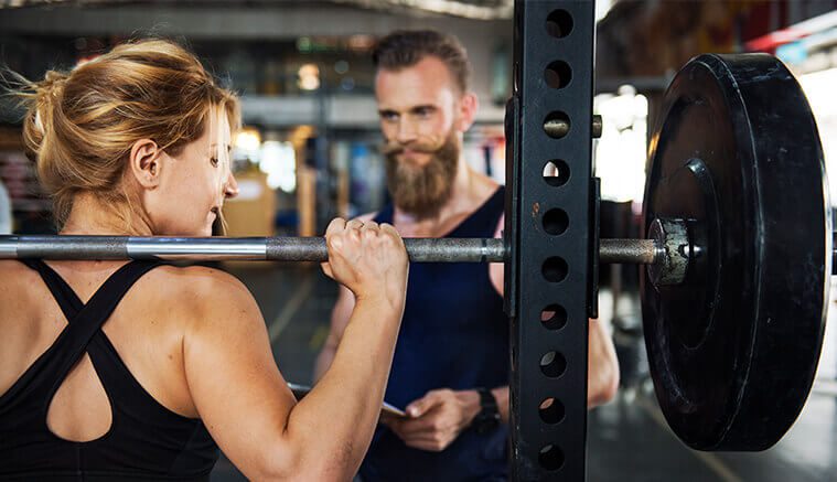 Male personal trainer assisting female client perform squats on a smith machine to highlight the importance of accident insurance