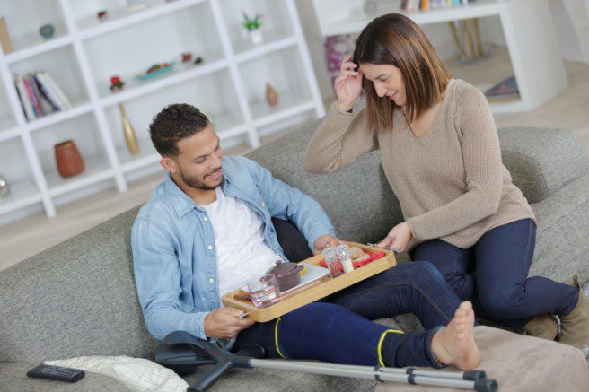 Female bringing food try to man of color with an injured leg sitting on sofa with crutches to promote disability insurance