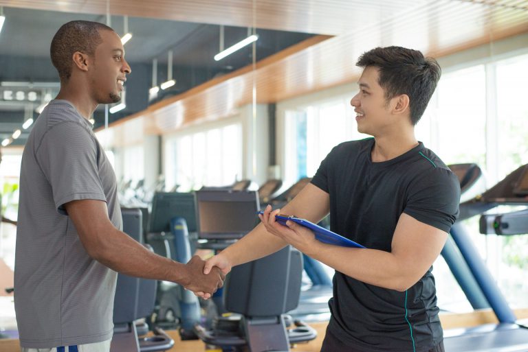Asian male personal trainer shaking hands with black male client in gym